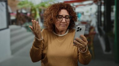 Şehir merkezinde akıllı telefon kullanan, orta yaşlı ve hayal kırıklığına uğramış bir kadın şehir ortamında, elinde kıvırcık esmer saçlı ve gözlüklü bir telefonla teknoloji etkileşimi tasvir ediyor.