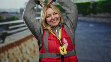 Şehir caddesinde kalp sembolü yapan, kırmızı yelek giyen ve açık havada gülümseyen sarı saçlı gönüllü bir kadın..