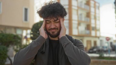 Güneşli bir günde, sakallı genç İspanyol bir adam açık havada, konut binalarının önünde dikiliyor, stresi ya da baş ağrısını şehir ortamında dile getiriyor..