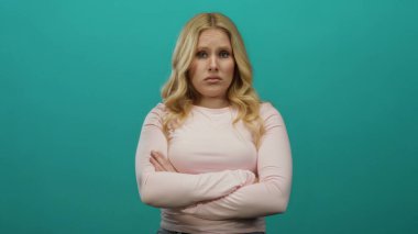 Sarı saçlı ve katlanmış kollu bir kadın pembe gömlek giyen canlı yeşil bir arka plana karşı güvenle duruyor..