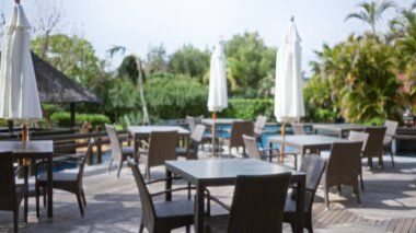 Boş hasır sandalyeler ve şemsiyelerle odaklanmış tropikal arka planı olan, ahşap bir verandaya kurulmuş, huzurlu bir tatil havası uyandıran bulanık bir açık hava tatil yeri sahnesi..