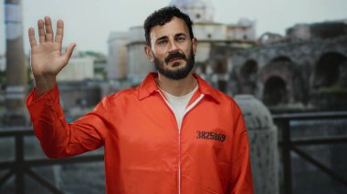 Parlak hapishane tulumu giymiş İspanyol bir adam, antik Roma arka planına karşı elini kaldırarak hapis ve kültürel birliktelik temalarını aktarıyor..