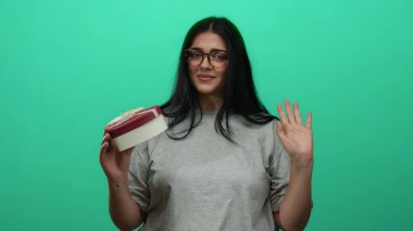 Gülümseyen bir kadın yeşil arka planda fiyonklu bir hediye kutusuyla uzun saç gözlükleriyle el sallayarak dostça bir karşılık veriyor.