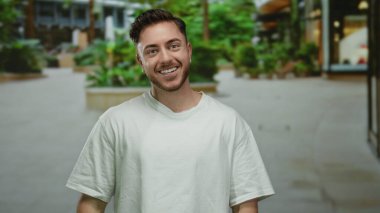 Şehir sokaklarında çapraz kolları olan genç bir adam, şehir yeşilliği ve modern mimarinin ortasında güvenle gülümsüyor..