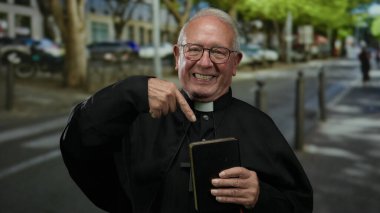 Başrahip bir şehir caddesinde dururken bir kitabı işaret ediyor. Çevresi kentsel unsurlar ve açık hava ortamında yeşilliklerle çevrili..