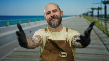 Deniz kenarındaki gezinti güvertesinde kolları açık gülümseyen, tulum ve eldiven giyen, dövmelerini gösteren, güneşli bir günde açık mavi gökyüzüne ve deniz zeminine karşı duran bir adam..