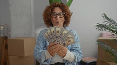 Yeni bir evde dolar banknotları gösteren bir kadın taşınan kutular ve bitkilerle çevrili, heyecan ve yeni bir başlangıç olasılığını somutlaştıran bir ev ortamında..