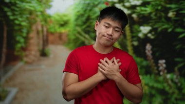 Kırmızı gömlekli genç, parkta göğsünü tutuyor, yeşil yemyeşil arka planda, duygularını ve iç mekanını gösteriyor..