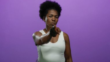 İzole edilmiş mor arkaplanı işaret eden bir kadın kararlı ifade ile beyaz kolsuz bluz giyiyor, güçlü ve kendinden emin bir varlık ve tutumu vurguluyor..