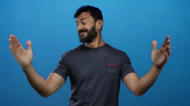 Sakallı İspanyol bir adam canlı mavi bir arkaplan önünde mutlu bir şekilde el hareketleriyle neşe ve açıklık ifade eden günlük koyu renk bir tişört giyiyor..