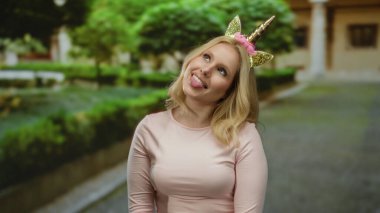 Sarışın bir kadın şehir sokaklarında tek boynuzlu bir bantla ağaçların ve binaların arasında komik bir ifadeyle komik bir poz vererek tuhaf bir sahne yaratıyor..