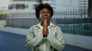 Şehir dışında açık havada dua eden gözlüklü bir kadın. Düşünceli bir ifadeyle kentsel çitlere karşı.