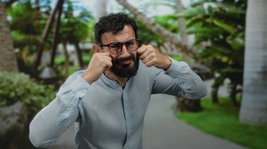 Gözlüklü İspanyol sakallı bir adam yeşil bir parkta, açık renk bir tişört giymiş ağaçlarla ağlıyor..