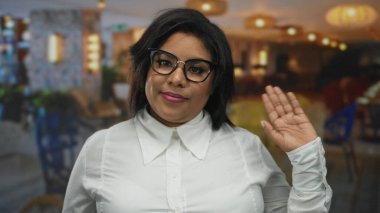 Restoran terasında gülümseyen gözlüklü ve beyaz gömlekli bir kadın şehrin arka planında neşeli bir güven gösterisi yapıyor..