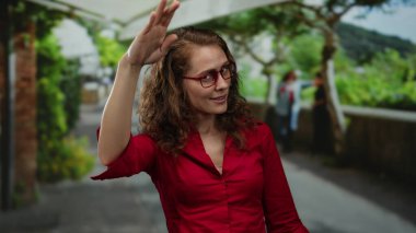 Parkta gözlüklü genç bir kadın, kırmızı bir tişört giyer ve güler yüzle dışarıda yeşillik ve yürüyüş arkaplanı olarak güler..