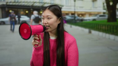 Pembe kazaklı uzun saçlı kadın şehir sokaklarında megafonla konuşuyor, şehir hayatı ve aktiviteleriyle çevrili, açık hava ortamında kendine güvenen iletişimi somutlaştırıyor..