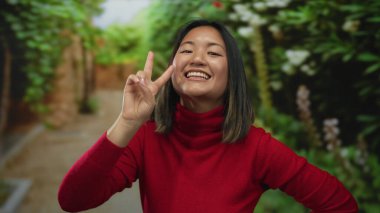 Kırmızı kazaklı genç bir kadın açık hava parkında gülümsüyor ve barış işareti gösteriyor.