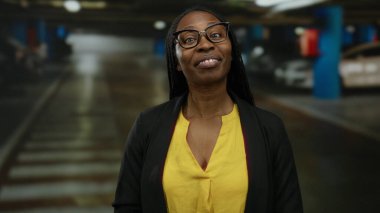 Kapalı otoparkta gülümseyen, sarı tişörtlü ve gözlüklü bir kadın. Arka planda bulanık arabalarla kendinden emin ve neşeli bir tavır sergiliyor..