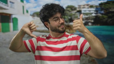 Sakallı, çizgili tişört giyen genç adam deniz kıyısında, bina ve okyanusun arka planında, açık havada karışık duygular olduğunu gösteren işaretler yapıyor..