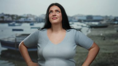 Sahilde kendine güvenen bir kadın, deniz kenarında düşünceli bir yüz ifadesiyle, açık mavi bir gömlek giymiş, elleri kalçasında ve arka planda botları olan..