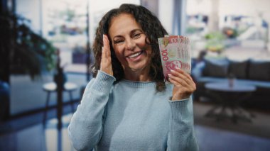Orta yaşlı bir kadın elinde yeni Zelanda banknotları tutarken gülümsüyor. Bir otel resepsiyonunda, zenginlik ve finansal başarı sergiliyor..