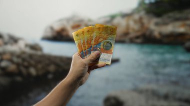 Sahilde İsviçre frangı banknotları tutan adam güneşli bir günde manzaralı su zeminine karşı para gösteriyor..