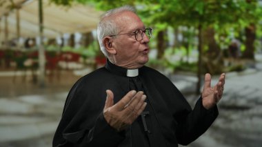 Kıdemli rahip gözlüklü bir adam sokakta jest yapıyor. Arka planda bulanık yeşil ağaçlar ve insanlar var. Huzurlu bir ruhaniyet hissi taşıyor..