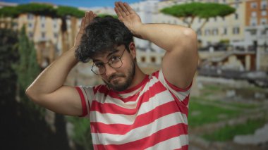 Kırmızı çizgili tişörtlü, sakallı genç İspanyol adam güneşli Roma harabelerinde kulak hareketi yaparak antik mimari ortamında neşeli ve güneşli bir anı yakalıyor..