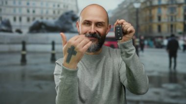 Dövmeli sakallı adam şehir merkezinin arka planında araba anahtarlarını gösteriyor. Canlı şehir dekorasyonu ve kültürel yaşam tarzında ulaşım araçlarının edinildiğini gösteriyor..