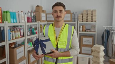 Yansıtıcı yelek giyen genç bir İspanyol, bağış merkezinde bir hediye kutusu tutuyor. Etrafında bir sürü malzeme var..