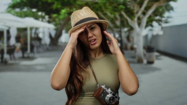 Güneşli bir günde başı ağrıyan bir kadın, şapka ve günlük giysiler giyiyor, turist kameraları asılı, arka planda ağaçlar ve bir kafe var..