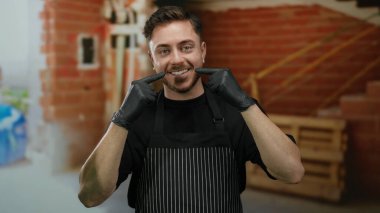 Sakallı, önlüklü ve eldivenli genç adam kısmen inşa edilmiş tuğla duvarlı bir evin içinde gülüşünü işaret ediyor..