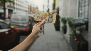 Şehir geçmişinde bulanık şehir geçmişiyle Kanada banknotları tutan adam, el ve para detaylarına odaklanmış açık havada para gösteriyor..