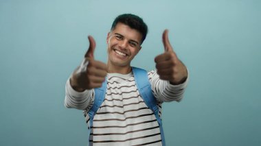 Mavi bir arkaplan önünde başparmağıyla gülümseyen genç adam, çizgili bir gömlek ve sırt çantası giyiyor, pozitifliğini ve güvenini ifade ediyor..