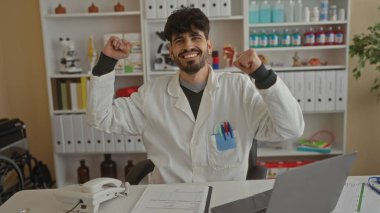 Klinikte gülümseyen genç bir adam laboratuvar önlüğü giyiyor ve ofis malzemeleriyle dolu bir masanın arkasında iyi organize edilmiş bir iş ortamında kutlama yapıyor.