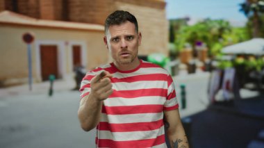 Kırmızı çizgili gömlekli genç adam, şehir sokağında kolunda dövmeleri olan etkileyici hareketler yapıyor.