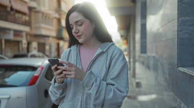 Şehir caddesinde açık havada akıllı telefon kullanan, arka planda park etmiş arabaları olan gri kapüşonlu bir kadın gülümsüyor..