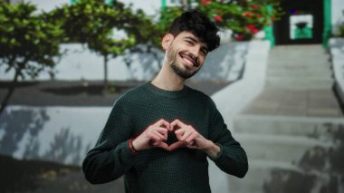 Bir kalp sembolü yapan genç bir adam şehir dışında renkli bir sanatsal arka plana karşı gülümseyen eller ile.