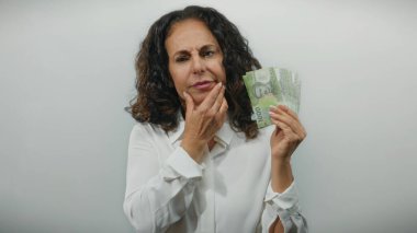 Orta yaşlı kadın elinde Şili pezosu olan beyaz bir arkaplan karşısında düşünceli bir jest yapıyor, finansal kararları ve fikirleri sembolize ediyor.