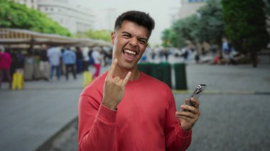 Kırmızı gömlekli genç adam, kalabalık bir şehir caddesinde insanlarla ve marketlerle dolu bir telefon tutarken sevinçle bir kaya işareti yapıyor..