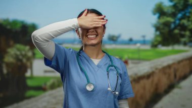 Mavi önlüklü kadın, güneşli bir park ortamında, steteskopunu ve neşeli yüz ifadesini göstererek, dışarıda neşeli bir anı temsil ediyor..