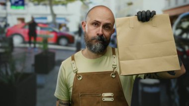 Şehirde sakallı bir adam, elinde kağıt torbayla önlük ve eldiven takıyor. Etrafı şehir çevresiyle çevrili..