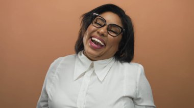 Gözlükleriyle turuncu arka planda gülümseyen bir kadın neşeli ifadesini ve büyük beden güzelliğini sergiliyor..