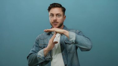 Sakallı genç adam izole edilmiş mavi arka plana karşı jest yapıyor, kot gömlek giyiyor, mola ya da duraklama ihtiyacını ifade ediyor, duygularını yüz ifadeleriyle ifade ediyor ve el işaretleriyle ifade ediyor..