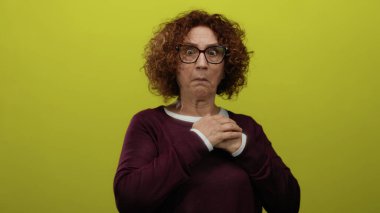 İspanyol orta yaşlı, kıvırcık saçlı, gözlüklü bir kadın canlı sarı izole bir arkaplan karşısında korkmuş görünüyor. İfadesinde duygu ve yoğunluk gösteriyor..