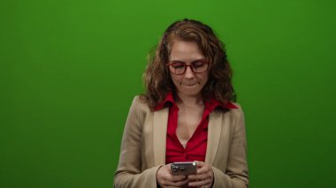 Akıllı telefon kullanan bir kadın, izole edilmiş yeşil arka planda kırmızı bluz ve kırmızı gözlüklü bej ceket giyiyor. Stüdyoda teknoloji ve iletişim kavramlarını tasvir ediyor..
