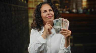 Kilisenin içinde İngiliz banknotları üzerinde kafa yoran, dini bir ortamda finansal tefekkür tasvir eden, merak ve parayla orta yaşlı bir kadını evin içinde ön plana çıkaran bir kadın..
