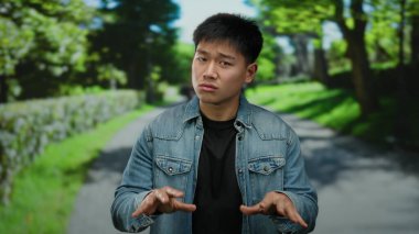 Genç adam, güneşli bir günde açık bir arka planda, yemyeşil bir parkta anlamlı jestler yapar.