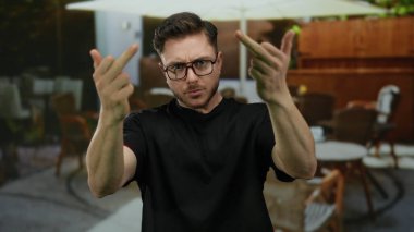Sakallı genç adam şehir kafeteryasının terasında kaba hareketler yapıyor. Etrafı masa ve sandalyelerle çevrili restoran atmosferiyle çevrili..