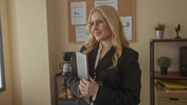 Modern ofiste gülümseyen bir kadın elinde tabletle, arka planda iç mekan bitkileri ve ofis malzemeleriyle profesyonelliği ve arkadaşlığı aktarıyor..